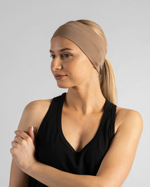 A studio portrait of a blonde woman in a three-quarter profile facing left, looking away from the camera against a plain light grey background. She is wearing a black sleeveless top and a beige merino ribbon headband, with her hair pulled back in a ponytail.