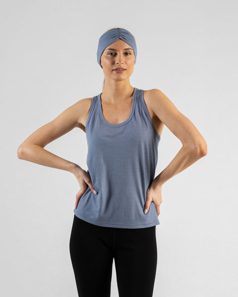 A studio portrait of a blonde woman standing with her hands on her hips, looking directly at the camera. She is wearing a dusty blue sleeveless top, black leggings, and a matching wide dusty blue headband with a gathered twist detail at the front, against a plain light grey background.