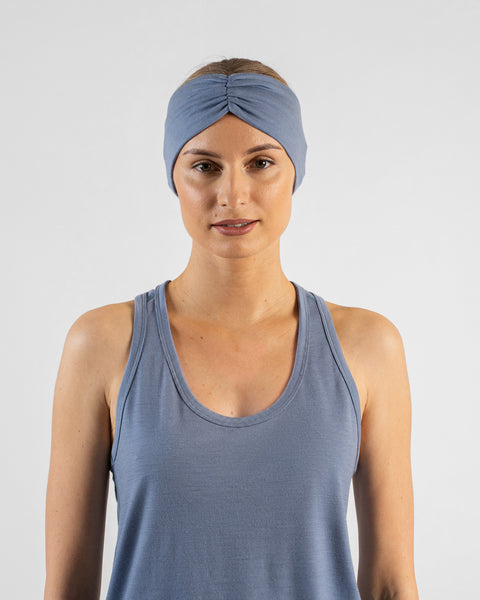 A close-up studio portrait of a blonde woman looking directly at the camera. She is wearing a dusty blue sleeveless top and a matching wide dusty blue headband with a gathered twist detail at the front, against a plain light grey background.