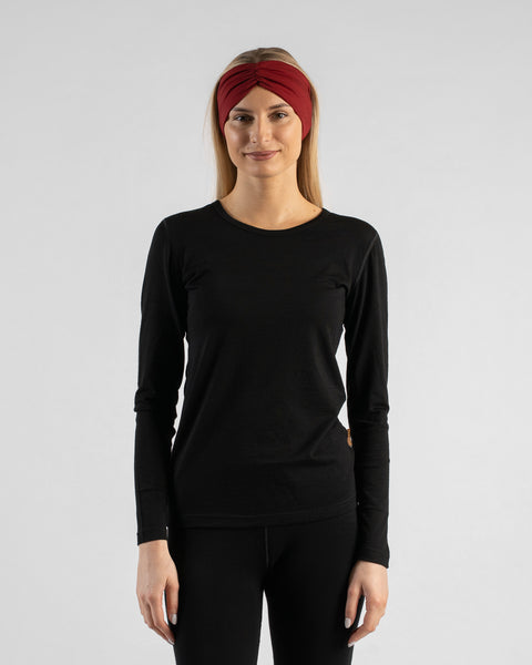A studio portrait of a blonde woman standing and looking directly at the camera with a soft smile against a plain light grey background. She is wearing a black long-sleeved shirt, black leggings, and a royal cherry deep red ribbon headband.