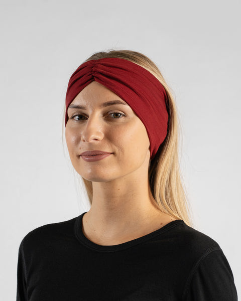 A close-up studio portrait of a blonde woman facing forward, looking directly at the camera with a soft smile against a plain light grey background. She is wearing a black long-sleeved shirt and a royal cherry deep red ribbon headband.