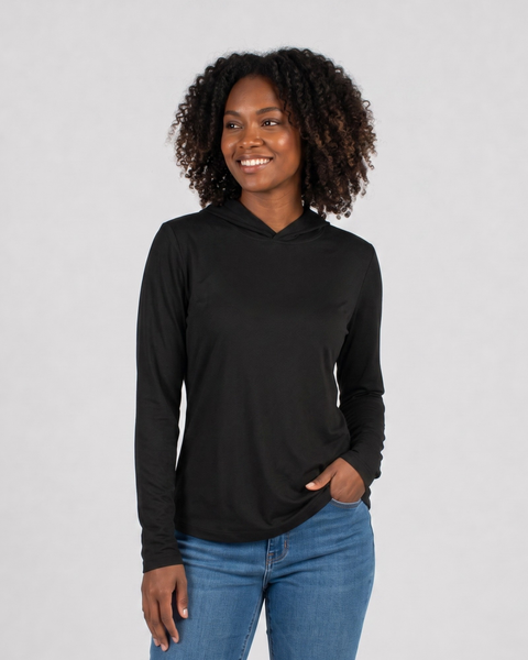 A smiling woman with curly hair looking to the side, wearing a black long-sleeved Women's Merino Sun Protective Hoodie with the hood worn down. She is wearing blue jeans and has one hand resting in her pocket against a light gray background.