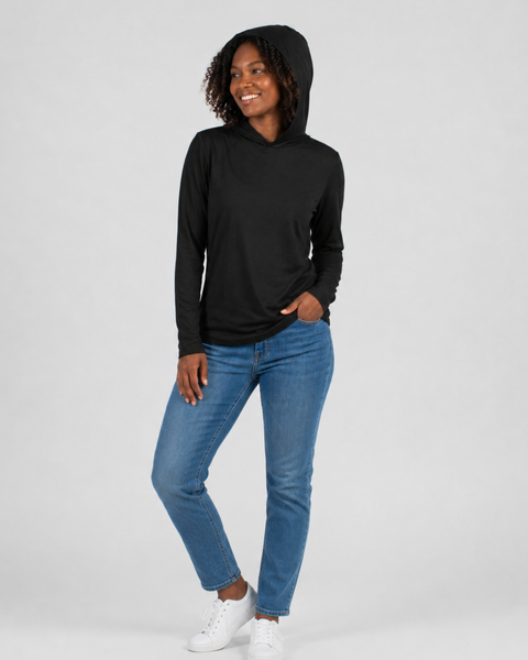 Full-body view of a smiling woman wearing a black long-sleeved Women's Merino Sun Protective Hoodie with the hood pulled up over her head. She is wearing blue jeans and white sneakers, with one hand in her pocket, standing against a light gray background.