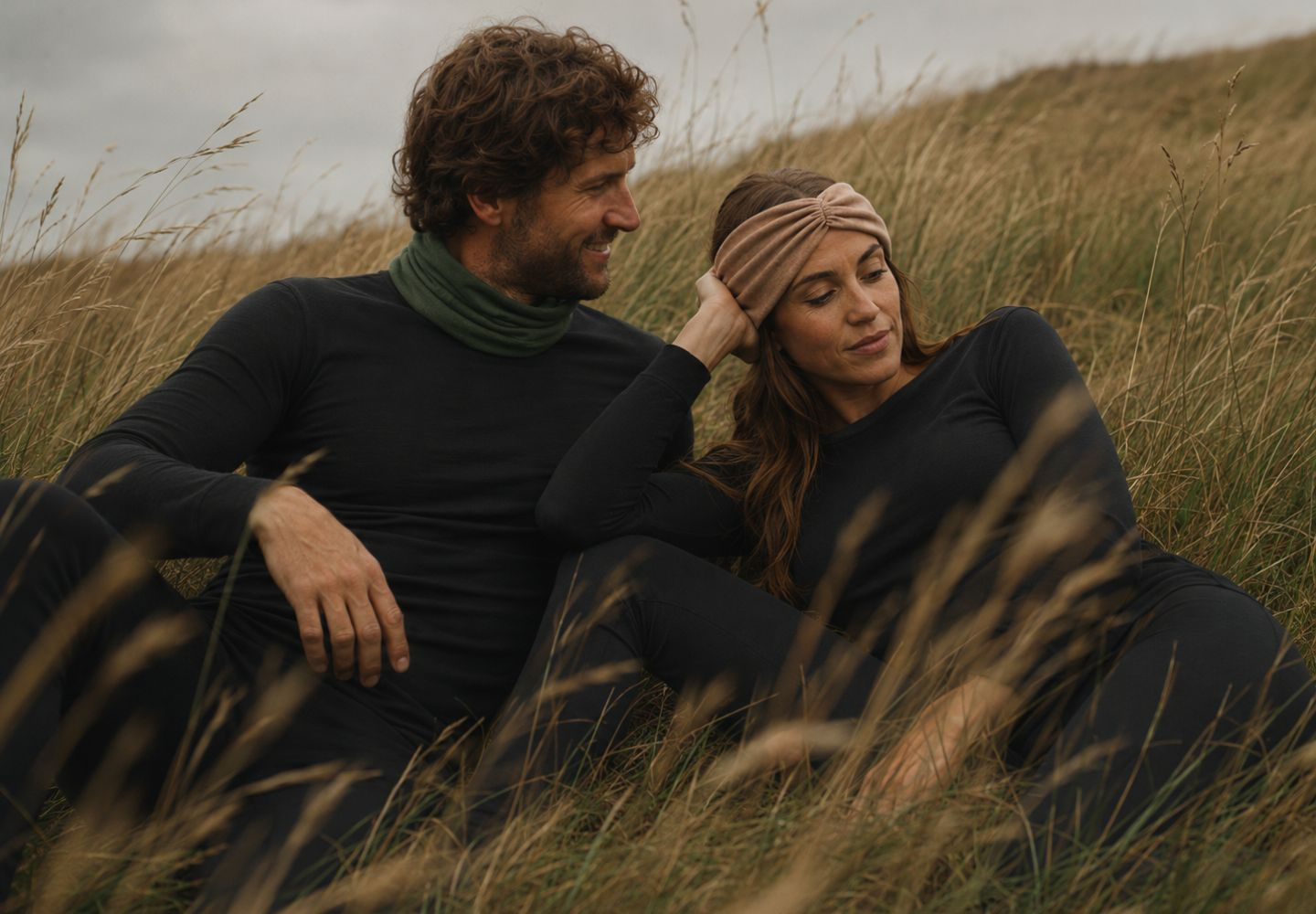 A smiling man in a black long-sleeved top and green neck gaiter sits beside a woman in a grassy field. The woman, wearing a matching black thermal top and a tan knit headband, looks softly downward as she rests her head near her hand.