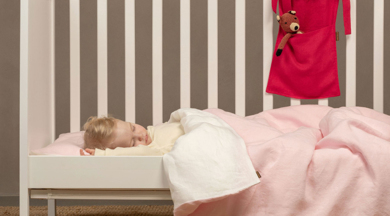 A landscape view of the sleeping toddler tucked under a pale pink duvet in a white wooden crib.
