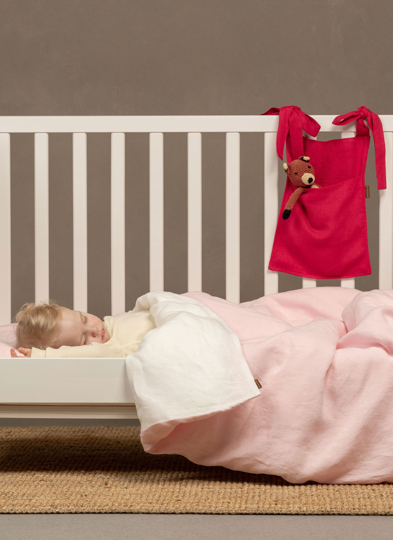 A vertical photo of a toddler sleeping peacefully in a white crib under a light pink blanket, with a red sleeping bag hanging nearby.