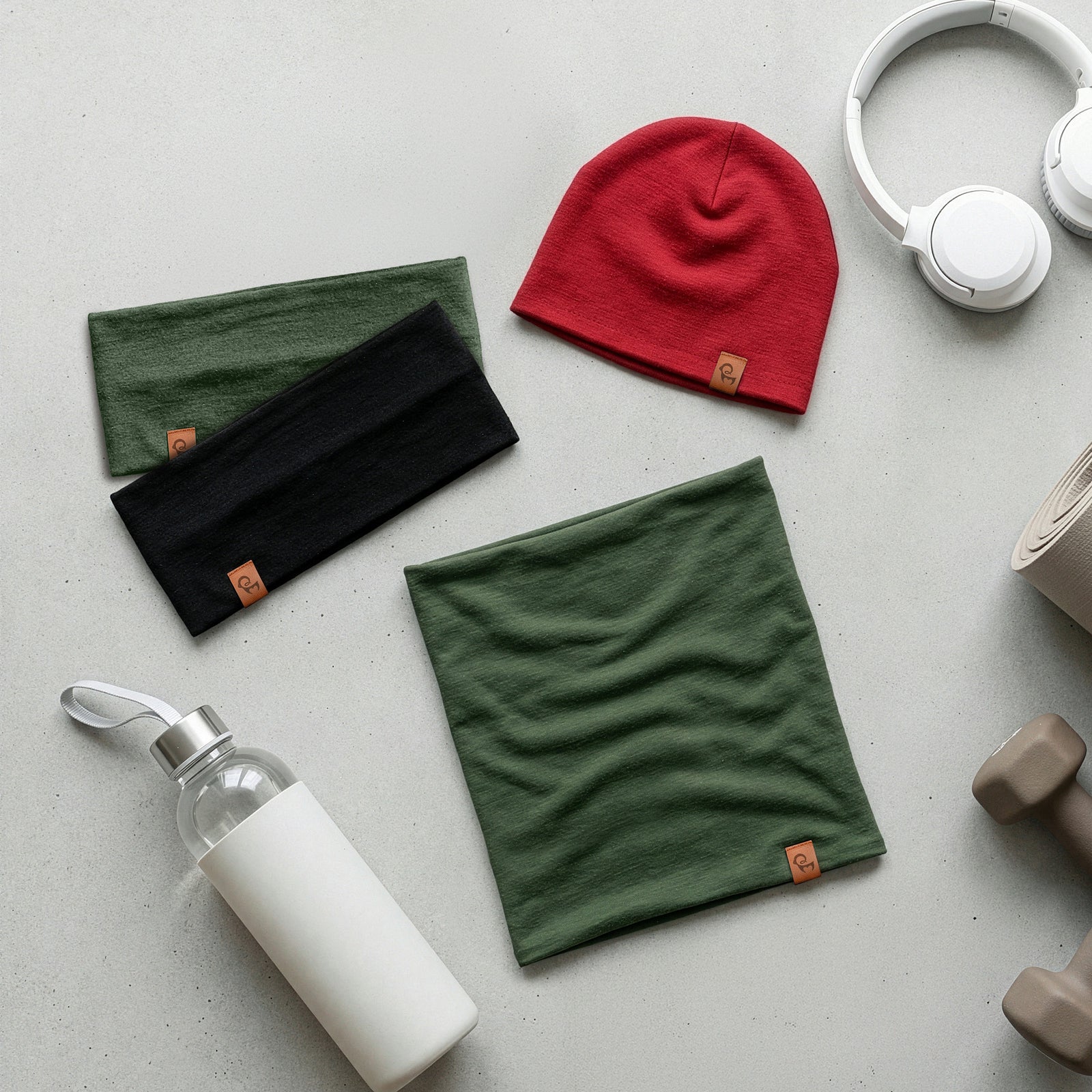 Flat lay of merino neck gaiters and a red beanie on a light surface with a water bottle and headphones.