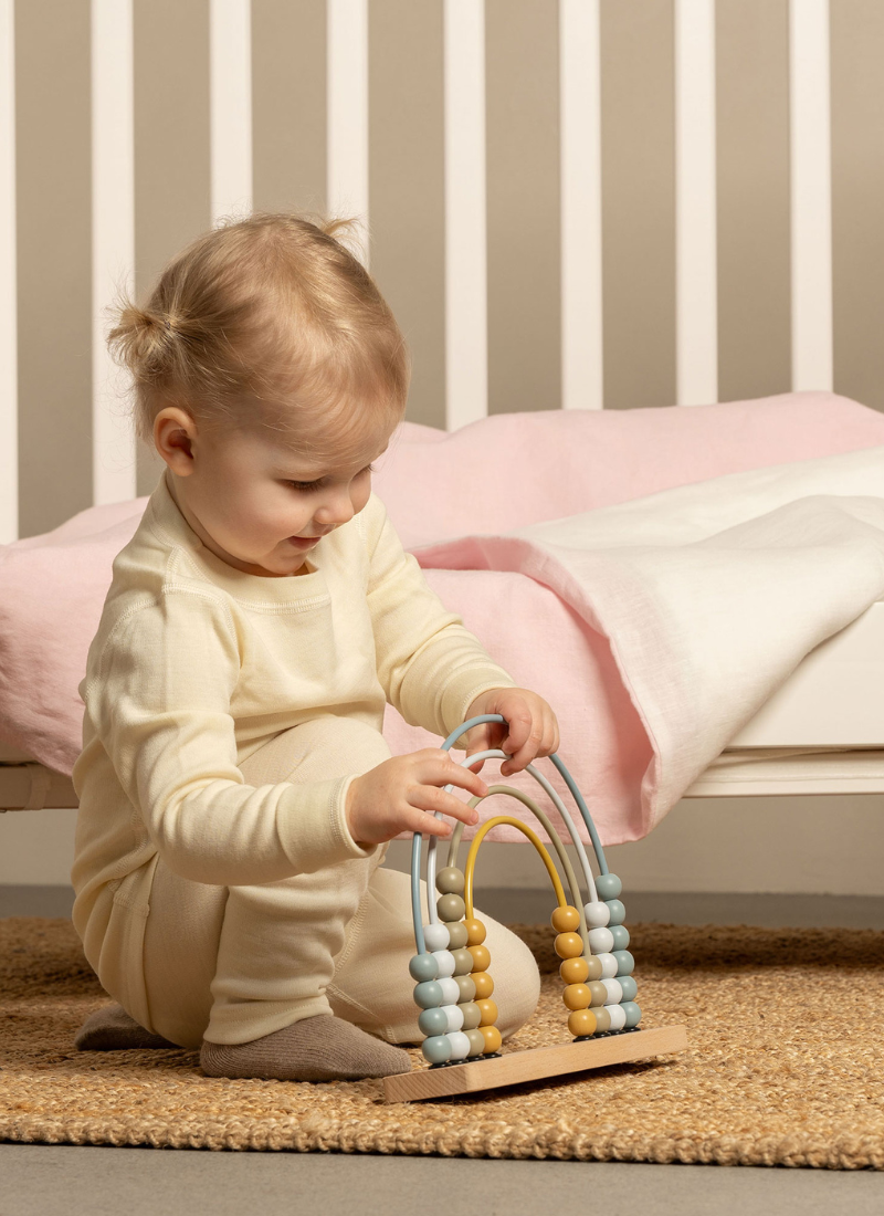 A portrait of a toddler in cream-colored knit pajamas sitting on a jute rug, playing with a wooden toy in front of a white crib.