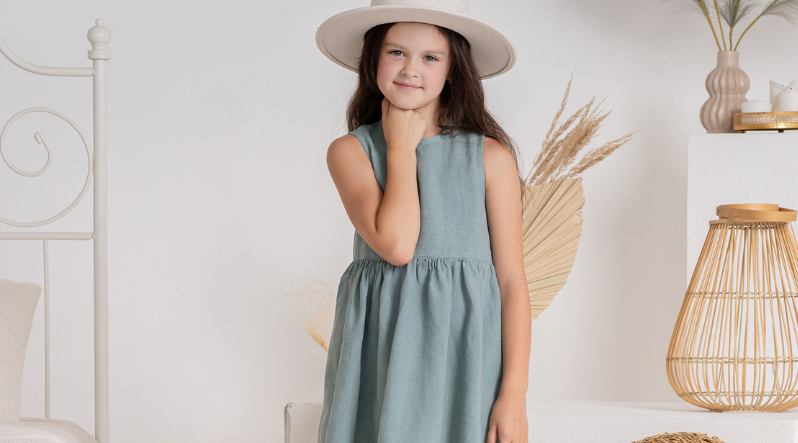 A young girl wearing a sleeveless light blue dress and a wide-brimmed straw hat looks thoughtfully toward the camera.