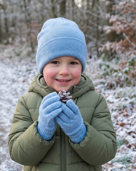 A young child smiling warmly while standing on a snowy path in a forest. The child is wearing an olive green hooded winter jacket, a light blue knit beanie, and matching light blue knit gloves, holding a small snow-covered pinecone with both hands.