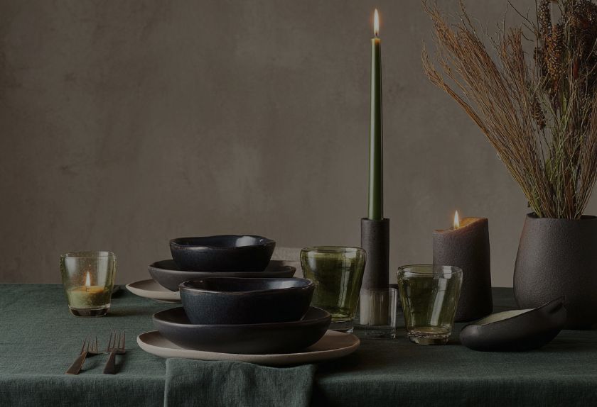 A moody table setting on a dark green tablecloth features stacked dark grey ceramic bowls, a tall green taper candle, and rustic botanical decor.