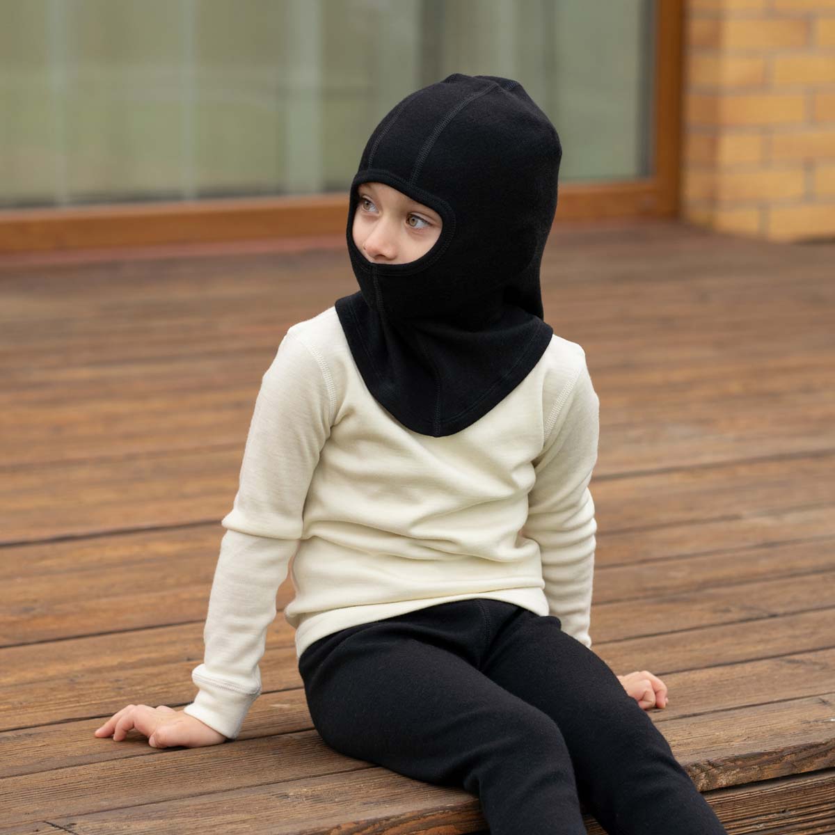 A young child wearing a royal cherry knit balaclava and a cream-colored shirt looks over their shoulder at the camera, with a background of blurred green foliage.