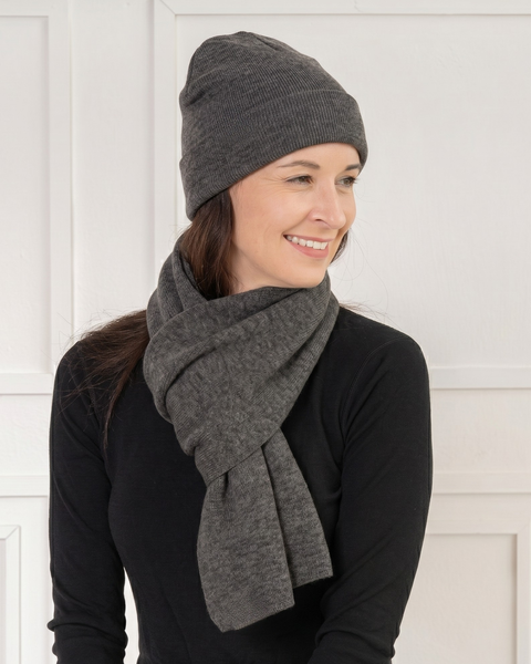 A smiling woman looking to the right, wearing a matching set of a dark grey knit beanie and a dark grey knit scarf wrapped once around her neck. She wears a black long-sleeved top against a background of a white wall with classic paneling.