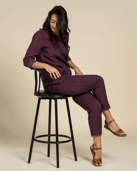 A woman sitting on a black stool wearing a shadow purple linen blouse with 3/4 sleeves and a high-rise waist, paired with matching linen pants.