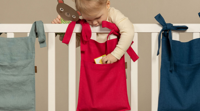 A landscape-oriented photo showing the teal, red, and navy baby sleeping bags neatly displayed on the rail of a white crib.